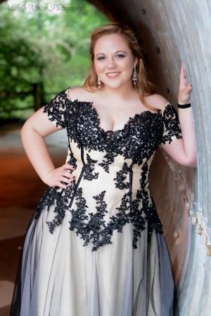 Fair skinned girl leaning against a curved cement wall in a pink dress with black overlay and lace trim with off the shoulder sleeves and a plunging neckline