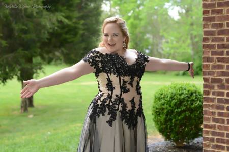 Fair skinned girl posing with arms out to the sides in a pink dress with black overlay and lace trim with off the shoulder sleeves and a plunging neckline