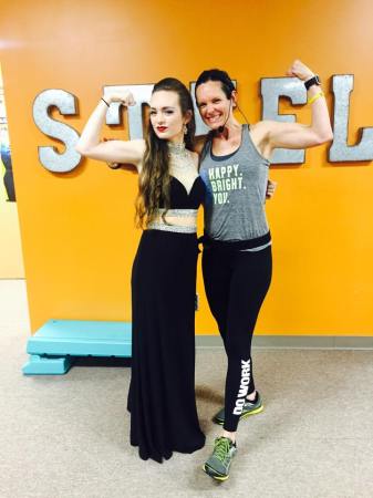 Fair skinned girl in a long black skirt with silver trim and a sweetheart neckline black top with silver trim. She is posing with her moth who is in gym clothes - black leggings and a grey tank top, both are flexing their biceps, they are in a gym.
